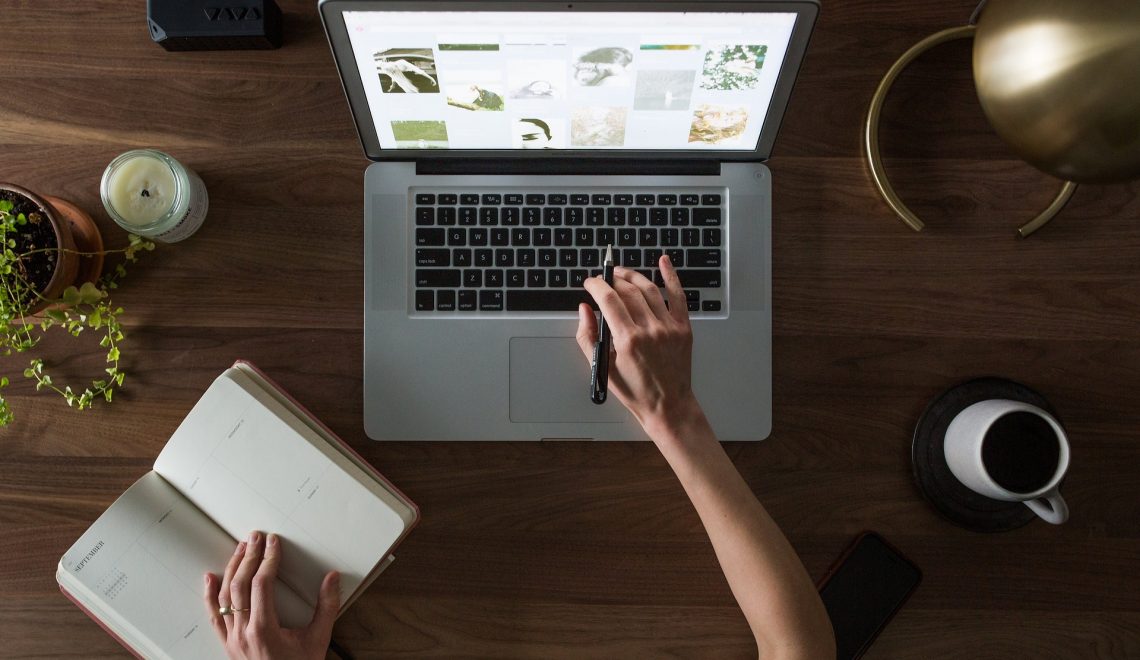 Visão aérea de uma pessoa, possivelmente um afiliado, sentada a uma mesa de madeira. A pessoa tem o dedo sobre o trackpad de um laptop, que está exibindo uma galeria de fotos. Há um caderno aberto, um smartphone, uma xícara de café, uma planta e uma vela na mesa.