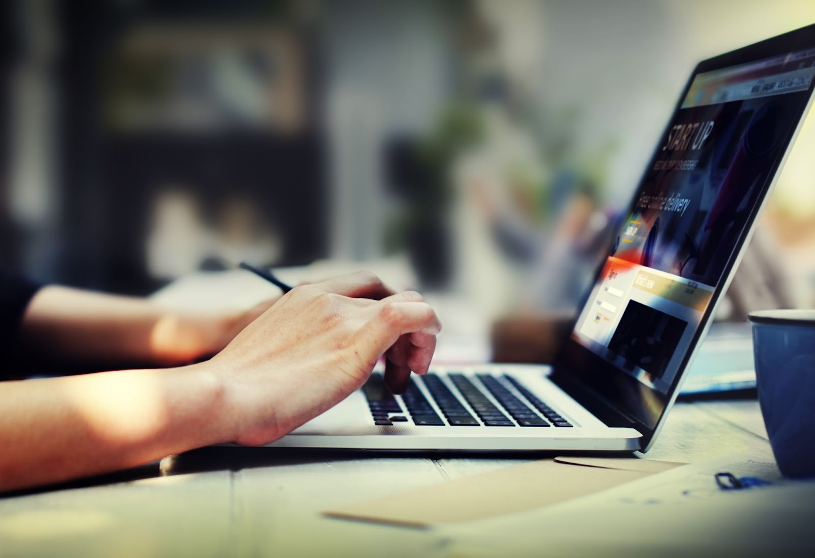 Visão lateral de uma pessoa, que atua como afiliado, digitando em um teclado de laptop. A tela do computador mostra um site com o título "START-UP", e a pessoa está trabalhando em uma mesa de madeira.
