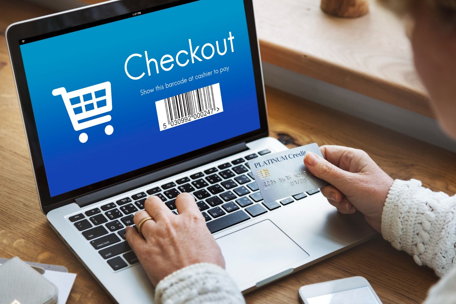 Uma pessoa está fazendo o checkout de compras online em um notebook, segurando um cartão de crédito. A tela do computador mostra a palavra "Checkout" e a imagem de um carrinho de compras com um código de barras.